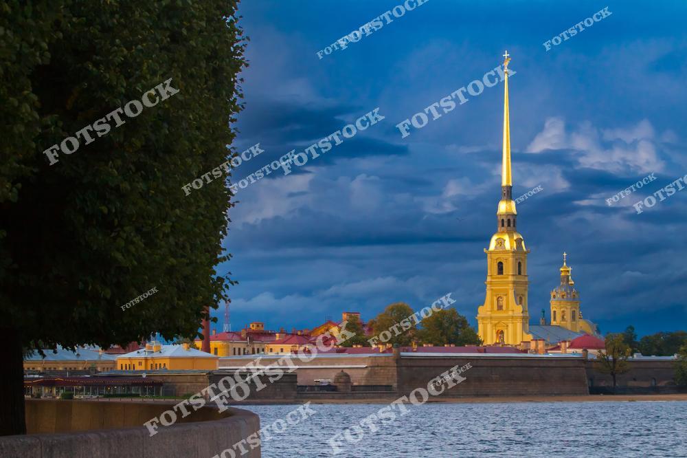 На фотографии изображена Петропавловская крепость, одна из главных достопримечательностей Санкт-Петербурга. Золотой шпиль собора святых Петра и Павла выделяется на фоне драматического вечернего неба. Крепость, расположенная на Заячьем острове, является историческим ядром города. Это место привлекает как туристов, так и местных жителей своей архитектурной и исторической значимостью. Вода Невы в переднем плане добавляет композиции спокойствия и величия.